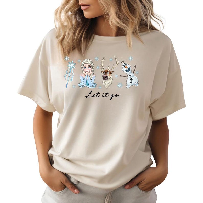 May include: Beige t-shirt featuring a graphic of Elsa, Olaf, Sven, and a snowflake wand. The phrase "Let it go" is written in black script below the characters. The shirt is a casual, short-sleeved style.