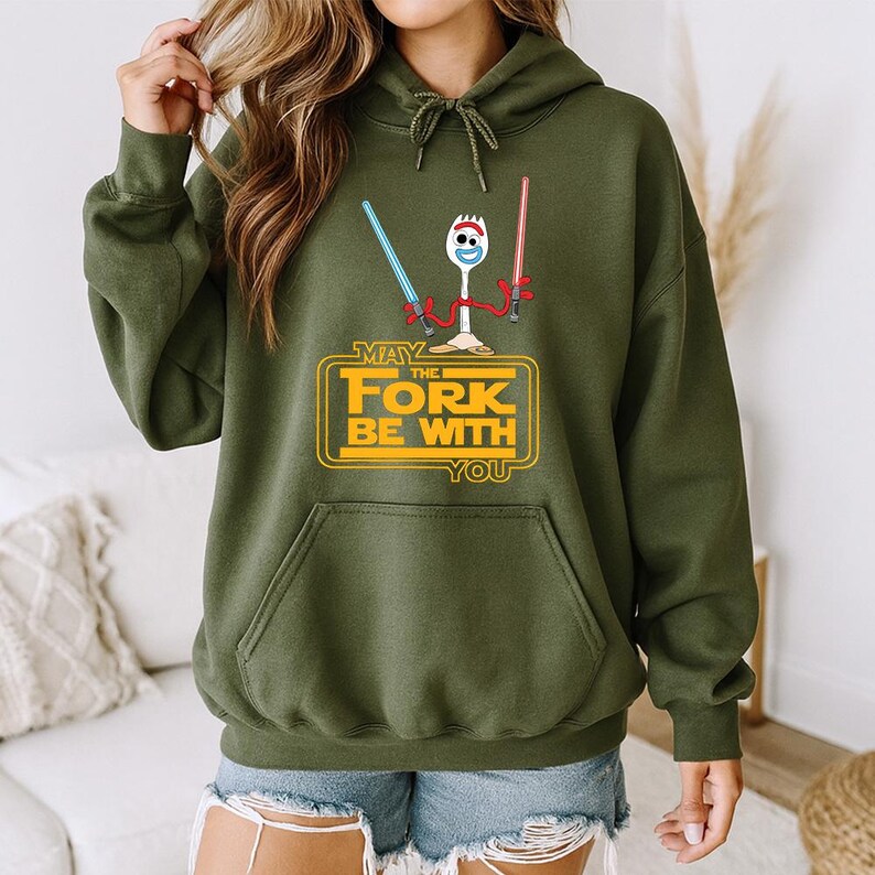 May include: An olive green hooded sweatshirt featuring a graphic of a white plastic spork character holding a blue and a red light saber. Below the character, the yellow text reads: MAY THE FORK BE WITH YOU, styled in a classic cinematic font.