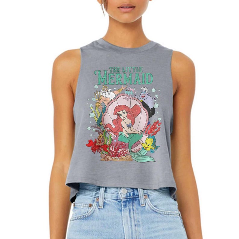 Puede incluir: Un top corto sin mangas de color gris jaspeado con un estampado gr&aacute;fico de los personajes de La Sirenita, incluyendo a Ariel sentada en una concha, el Rey Trit&oacute;n, &Uacute;rsula, Sebasti&aacute;n y Flounder, con el texto The Little Mermaid impreso en verde.