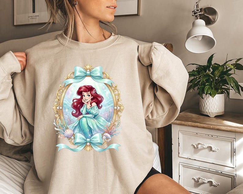 May include: Beige sweatshirt featuring a framed illustration of a mermaid with red hair and a teal dress. The illustration is surrounded by a gold frame and teal bows. The mermaid is sitting in front of a blue and white background.