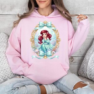 Puede incluir: Sudadera con capucha rosa claro con un dise&ntilde;o de sirena de dibujos animados en un marco dorado, adornada con lazos verde azulado. La sirena tiene el pelo rojo y un vestido verde azulado. La sudadera tiene un bolsillo delantero y una capucha.