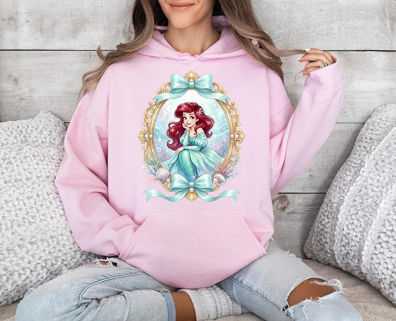 Puede incluir: Sudadera con capucha rosa claro con un dise&ntilde;o de sirena de dibujos animados en un marco dorado, adornada con lazos verde azulado. La sirena tiene el pelo rojo y un vestido verde azulado. La sudadera tiene un bolsillo delantero y una capucha.