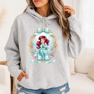 Puede incluir: Sudadera con capucha gris claro con una ilustraci&oacute;n enmarcada de una sirena con cabello rojo y un vestido turquesa. El marco dorado tiene lazos turquesas. La sirena est&aacute; rodeada de conchas marinas y plantas acu&aacute;ticas.