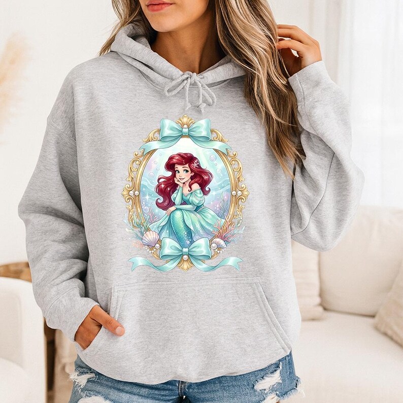 Puede incluir: Sudadera con capucha gris claro con una ilustraci&oacute;n enmarcada de una sirena con cabello rojo y un vestido turquesa. El marco dorado tiene lazos turquesas. La sirena est&aacute; rodeada de conchas marinas y plantas acu&aacute;ticas.