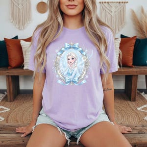 May include: A light purple t-shirt featuring a cartoon image of a princess in a decorative oval frame with blue bows. The princess has blonde hair and is wearing a blue dress. The shirt is paired with denim shorts.