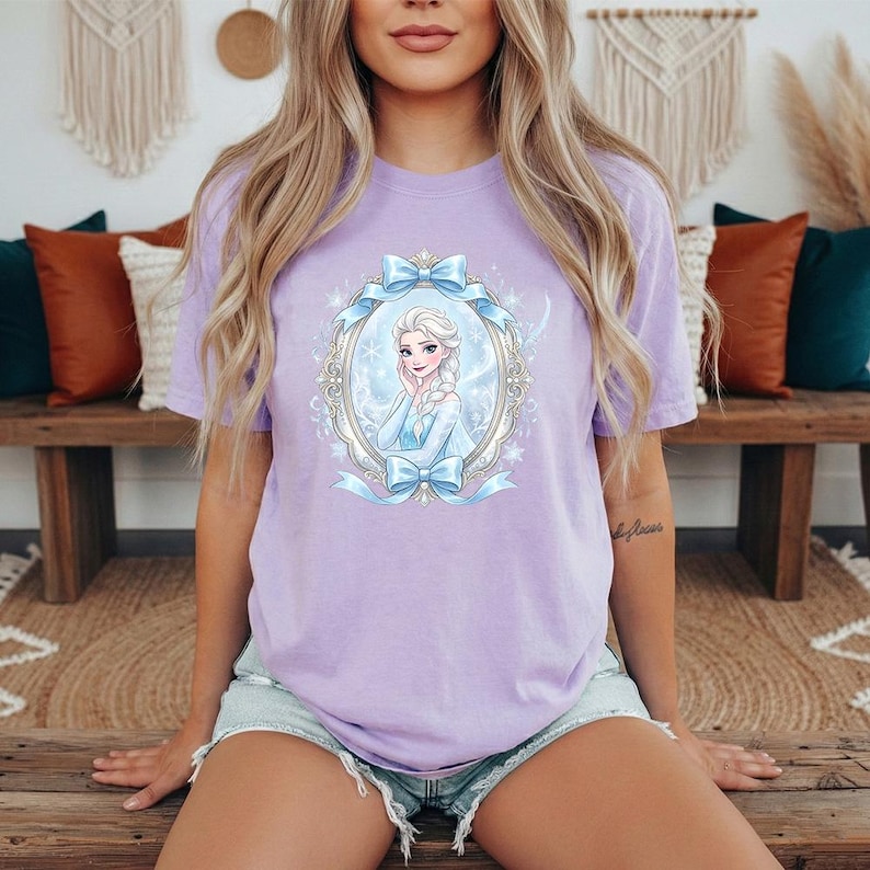 May include: A light purple t-shirt featuring a cartoon image of a princess in a decorative oval frame with blue bows. The princess has blonde hair and is wearing a blue dress. The shirt is paired with denim shorts.
