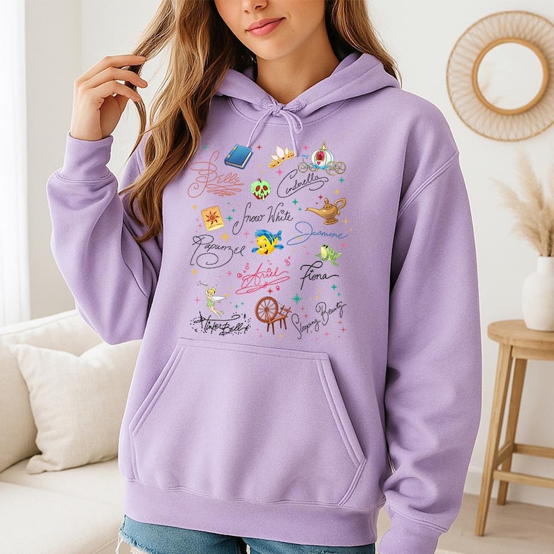 May include: A light purple hooded sweatshirt featuring a graphic print of various fairy tale character names in cursive script, including Belle, Cinderella, Snow White, Jasmine, Rapunzel, Ariel, Fiona, Tinker Bell, and Sleeping Beauty, with icons.