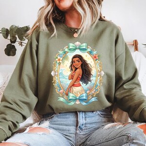 May include: Olive green sweatshirt featuring a framed illustration of a cartoon woman with long dark hair, wearing a red and white top and skirt. The frame includes a blue bow, flowers, and seashells. The background shows a beach scene.