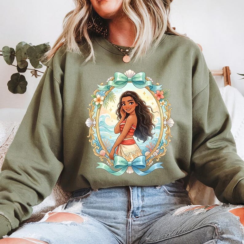 May include: Olive green sweatshirt featuring a framed illustration of a cartoon woman with long dark hair, wearing a red and white top and skirt. The frame includes a blue bow, flowers, and seashells. The background shows a beach scene.