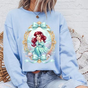 May include: Light blue sweatshirt featuring a framed illustration of a mermaid with red hair and a teal dress. The illustration is surrounded by gold trim, bows, and seashells. The sweatshirt is a solid light blue color.