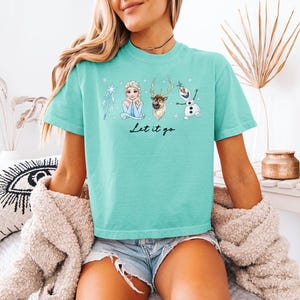 May include: A turquoise t-shirt featuring illustrations of characters from the movie "Frozen", including Elsa, a reindeer, and Olaf the snowman. The phrase "Let it go" is written in a script font below the characters. The shirt is a short-sleeved, crew neck style.