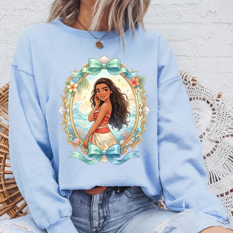 May include: Light blue sweatshirt featuring a framed illustration of a woman with long dark hair, wearing a red and white top and a white skirt. The frame is gold with floral and seashell accents, and teal bows.
