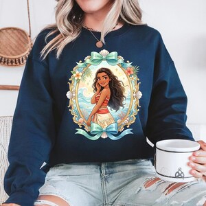 May include: Navy blue sweatshirt featuring a colorful illustration of a woman in a red and white top and a white skirt, framed by a gold oval with floral and seashell accents. The design includes a light blue bow at the top and bottom. The sweatshirt is a crew neck style.