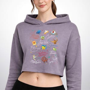 May include: A mauve cropped hoodie featuring a graphic print of various animated character names and icons, including a blue book, a golden lamp, a green apple, a spinning wheel, and small colorful stars scattered across the center chest area.