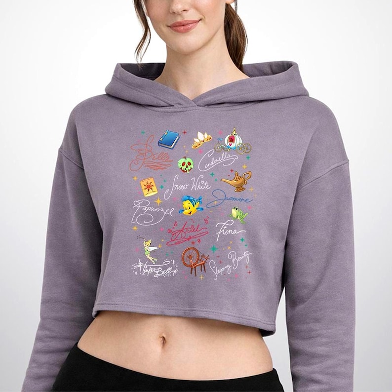 May include: A mauve cropped hoodie featuring a graphic print of various animated character names and icons, including a blue book, a golden lamp, a green apple, a spinning wheel, and small colorful stars scattered across the center chest area.