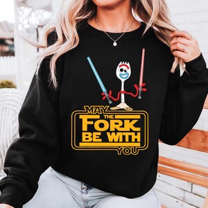 May include: A black long-sleeve sweatshirt featuring a graphic of a white spork character with googly eyes holding a blue and a red lightsaber. Below the character, yellow text reads "MAY THE FORK BE WITH YOU" inside a rectangular border.