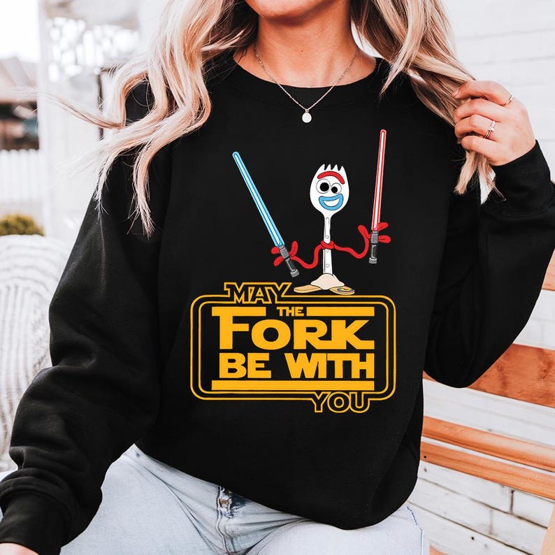 May include: A black long-sleeve sweatshirt featuring a graphic of a white spork character with googly eyes holding a blue and a red lightsaber. Below the character, yellow text reads "MAY THE FORK BE WITH YOU" inside a rectangular border.