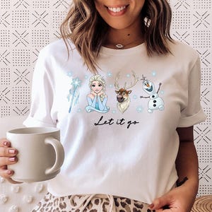 May include: White t-shirt featuring characters from the animated film "Frozen." The design includes Elsa, Olaf, Sven, and a snowflake-themed wand. The phrase "Let it go" is written in black script below the characters.