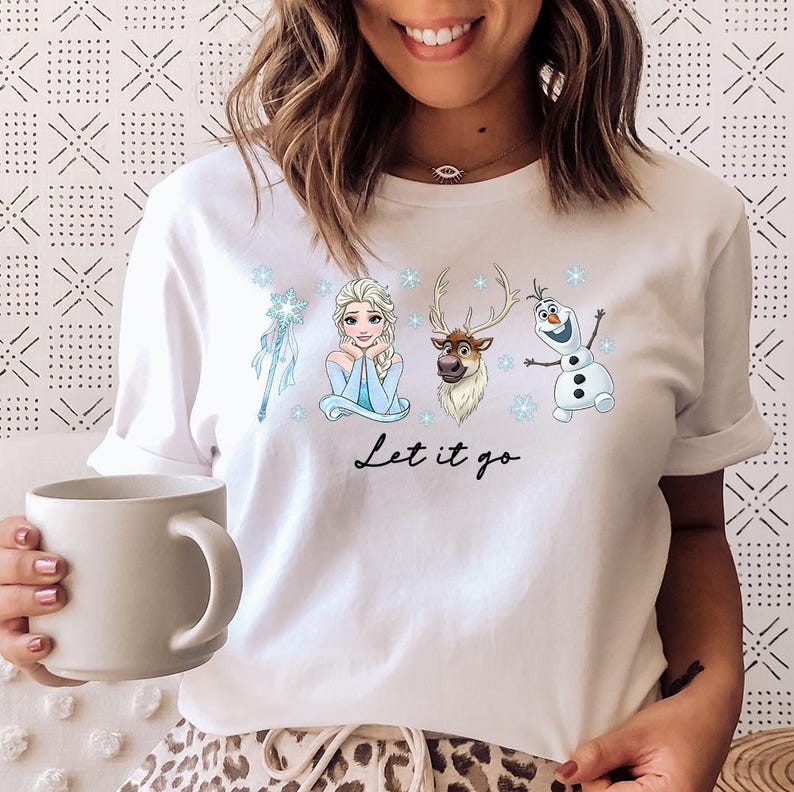 May include: White t-shirt featuring characters from the animated film "Frozen." The design includes Elsa, Olaf, Sven, and a snowflake-themed wand. The phrase "Let it go" is written in black script below the characters.