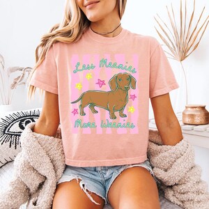 May include: A peach-coloured short-sleeve t-shirt featuring a graphic of a brown dachshund dog. The text "Less Meanies More Weenies" is printed in teal above and below the dog, surrounded by small pink and yellow star illustrations.