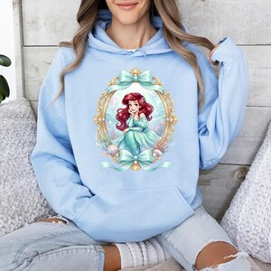 Puede incluir: Sudadera con capucha azul claro con una ilustraci&oacute;n enmarcada de una sirena pelirroja con un vestido turquesa. El marco es dorado con lazos turquesa. La sudadera tiene una capucha con cord&oacute;n y un bolsillo delantero.