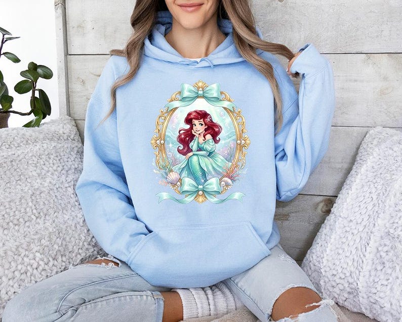 Puede incluir: Sudadera con capucha azul claro con una ilustraci&oacute;n enmarcada de una sirena pelirroja con un vestido turquesa. El marco es dorado con lazos turquesa. La sudadera tiene una capucha con cord&oacute;n y un bolsillo delantero.