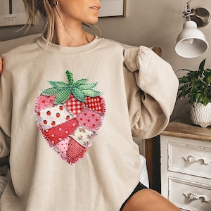 May include: Beige sweatshirt featuring a large, patchwork strawberry design. The strawberry is composed of various red and pink fabrics, including polka dots, hearts, and gingham. The stem and leaves are green. A casual, comfortable top.