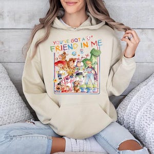 May include: Beige hoodie featuring a colorful graphic of characters from the animated film "Toy Story." The graphic includes the text "YOU'VE GOT A FRIEND IN ME." The hoodie has a front pocket and a drawstring hood.