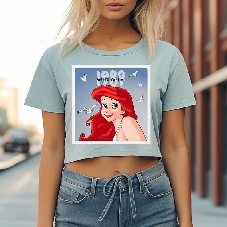 May include: Light blue crop top featuring a square graphic of Ariel from The Little Mermaid, with the text "1989 Ariel's Version". The graphic shows Ariel with red hair and seagulls. The shirt is paired with blue jeans.