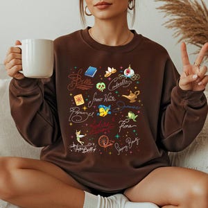May include: A dark brown crewneck sweatshirt featuring a colorful graphic print of various fairy tale icons and character names including Belle, Cinderella, Snow White, Jasmine, Rapunzel, Fiona, Ariel, Tinker Bell, and Sleeping Beauty.