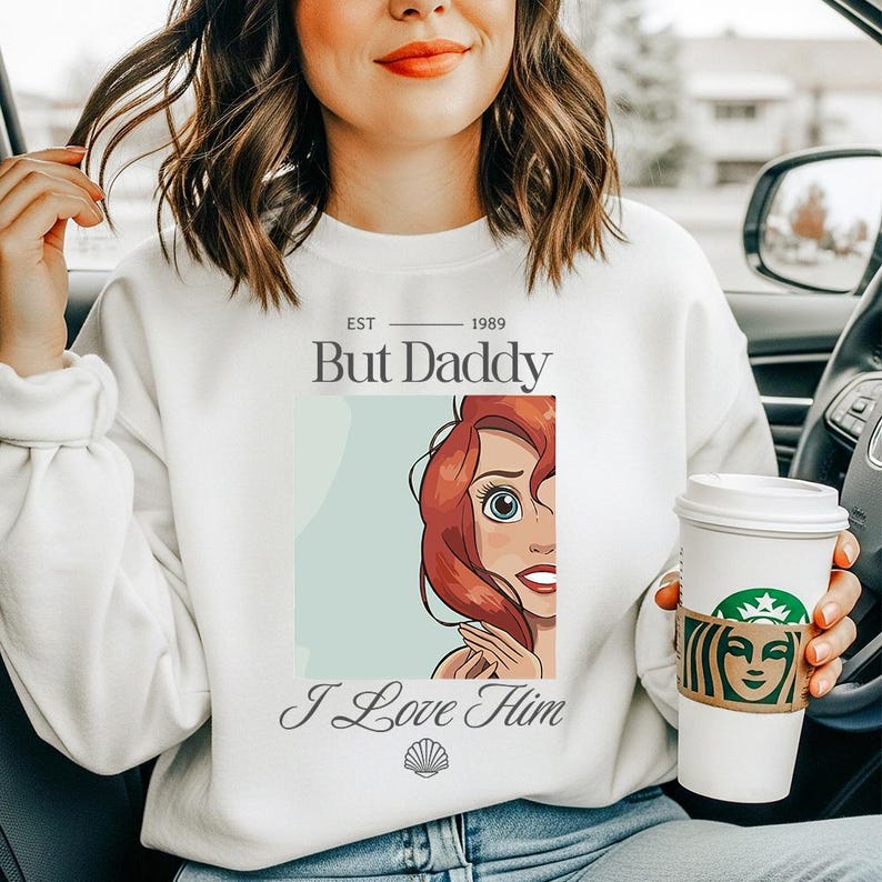 May include: White sweatshirt with the text "But Daddy" above a cartoon image of a red-haired character. Below the image is the text "I Love Him" and a seashell graphic. The sweatshirt also has the text "EST 1989".
