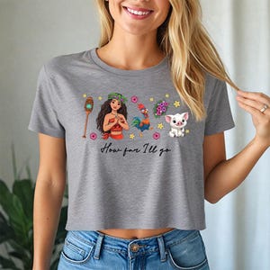 May include: A grey crop top with a vibrant graphic design featuring characters and the text "How far I'll go". The design includes a character with a flower crown, a rooster, a pig, and other elements. The shirt is made of a soft material.