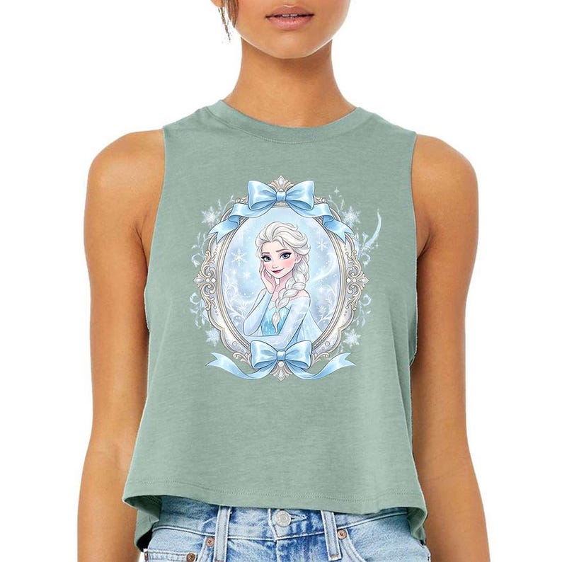 Pu&ograve; includere: Top corto verde salvia con un'illustrazione incorniciata di una principessa con un vestito blu e treccia bionda. La cornice &egrave; decorata con fiocchi blu e fiocchi di neve. Il top &egrave; senza maniche e in materiale morbido.