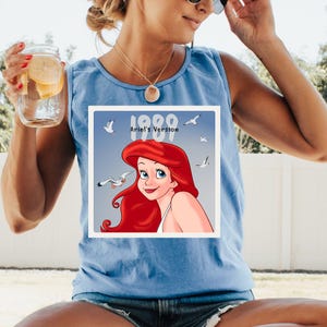 May include: Light blue tank top featuring a square graphic of Ariel from The Little Mermaid, with red hair and blue eyes, surrounded by seagulls. The graphic includes the text "1989 Ariel's Version".