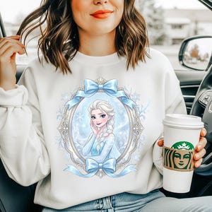 K&ouml;nnte beinhalten: Wei&szlig;es Sweatshirt mit einem Cartoon-Portr&auml;t einer Figur in einem blauen Kleid und einem Zopf, umrahmt von blauen Schleifen. Die Person, die das Sweatshirt tr&auml;gt, sitzt in einem Auto und h&auml;lt einen Kaffeebecher.