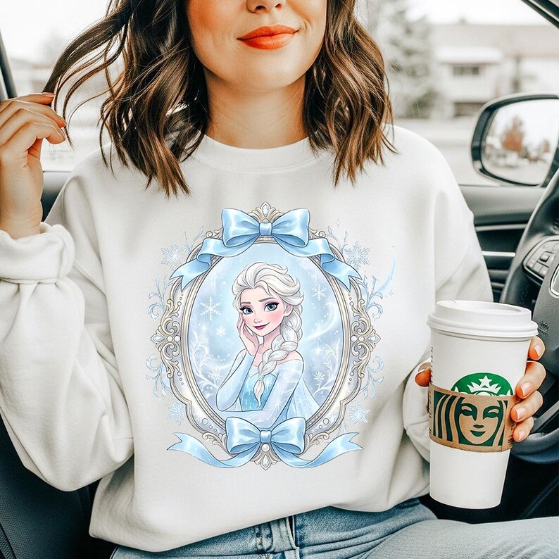K&ouml;nnte beinhalten: Wei&szlig;es Sweatshirt mit einem Cartoon-Portr&auml;t einer Figur in einem blauen Kleid und einem Zopf, umrahmt von blauen Schleifen. Die Person, die das Sweatshirt tr&auml;gt, sitzt in einem Auto und h&auml;lt einen Kaffeebecher.