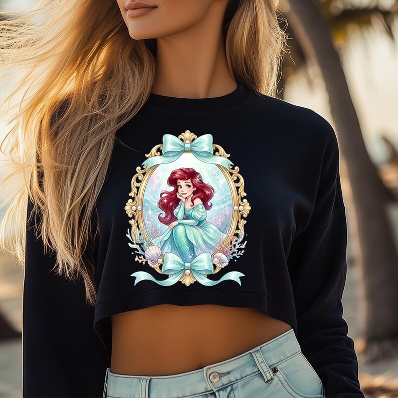 Puede incluir: Sudadera corta negra con una ilustraci&oacute;n enmarcada de una sirena con cabello rojo y un vestido turquesa. El marco es dorado con lazos y conchas marinas turquesas. Una parte superior informal.