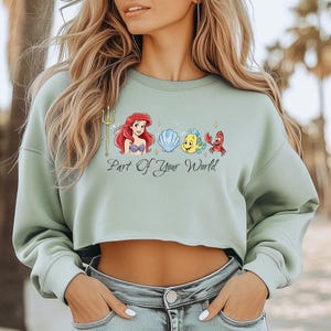 May include: A pale green cropped sweatshirt with a graphic of Ariel, Flounder, Sebastian, and a seashell. The words "Part of Your World" are printed beneath the characters. The sweatshirt has long sleeves and a round neck.