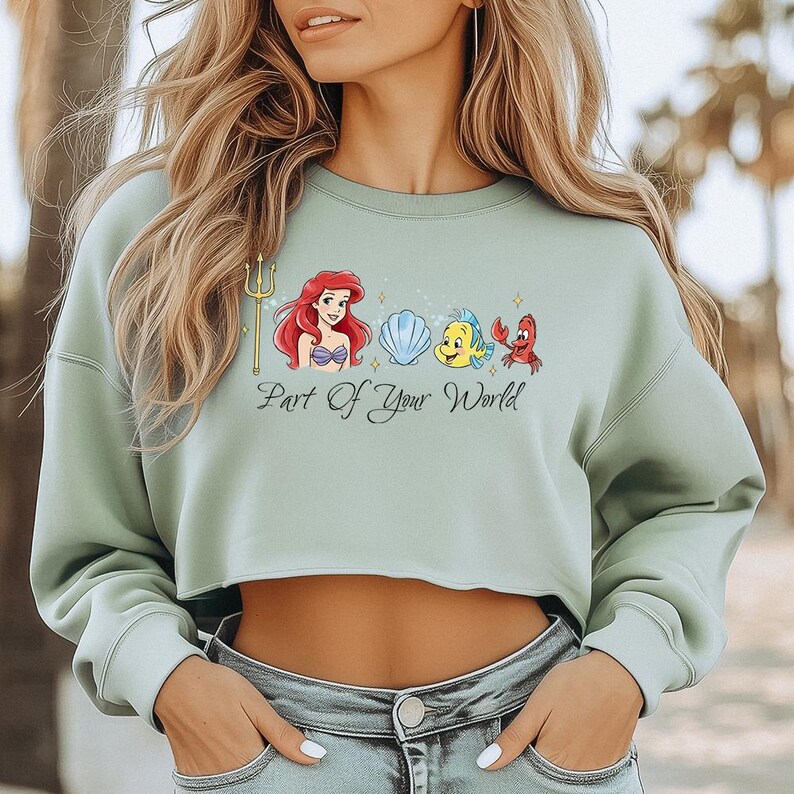 May include: A pale green cropped sweatshirt with a graphic of Ariel, Flounder, Sebastian, and a seashell. The words "Part of Your World" are printed beneath the characters. The sweatshirt has long sleeves and a round neck.