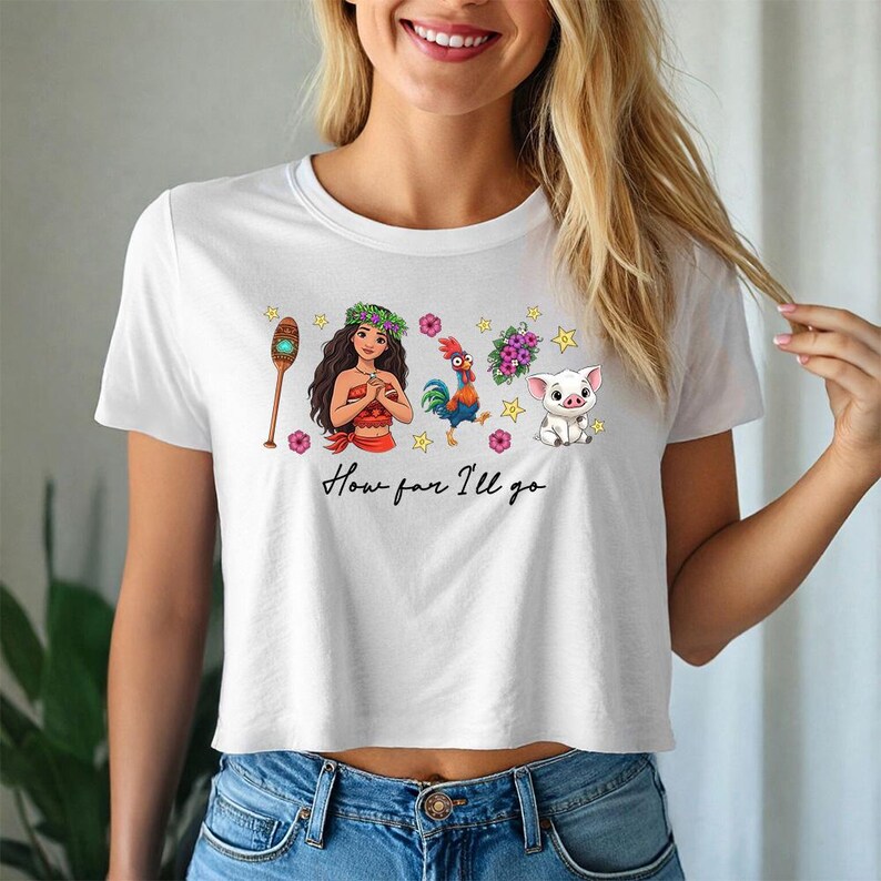 May include: White crop top with a colorful graphic design featuring characters and the text "How far I'll go". The design includes a cartoon woman, rooster, pig, and other elements. The shirt is a casual style.