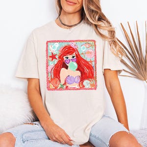 May include: Cream-colored t-shirt featuring a colorful graphic of a mermaid with red hair, heart-shaped sunglasses, and a bubblegum bubble. The graphic includes seashells, starfish, and fish. The mermaid is wearing a purple seashell bra.