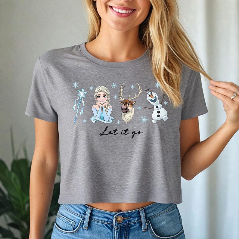 May include: Gray crop top featuring Elsa, Olaf, and Sven from Frozen, with snowflakes and the phrase "Let it go." The shirt has a crew neck and short sleeves. The graphic is in a cartoon style.