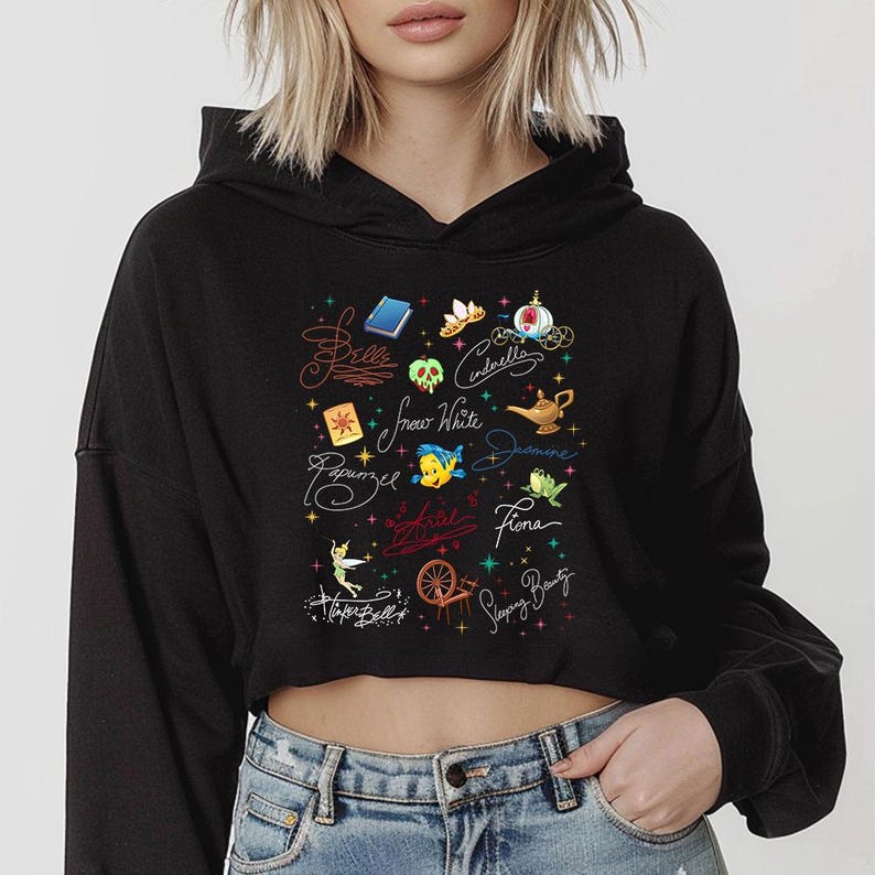 May include: A black cropped hoodie featuring a graphic print of various fairy tale icons and character names including Belle, Cinderella, Snow White, Jasmine, Rapunzel, Ariel, Fiona, Tinker Bell, and Sleeping Beauty, accented with colorful stars.