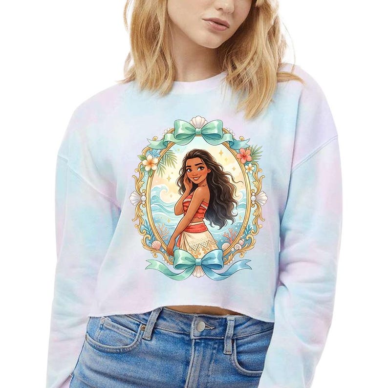 Peut inclure: Un sweat-shirt court tie-dye avec une illustration encadr&eacute;e d'une femme de dessin anim&eacute; aux longs cheveux noirs, v&ecirc;tue d'une tenue rouge et blanche. Le cadre est d&eacute;cor&eacute; de fleurs, de coquillages et de n&oelig;uds bleu sarcelle. Le fond est un tie-dye bleu clair, rose et violet.