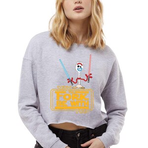 May include: A light gray cropped sweatshirt featuring a graphic of a white spork character with red pipe cleaner arms holding a blue and a red lightsaber. Below the character, the yellow text reads MAY THE FORK BE WITH YOU in a stylized font.