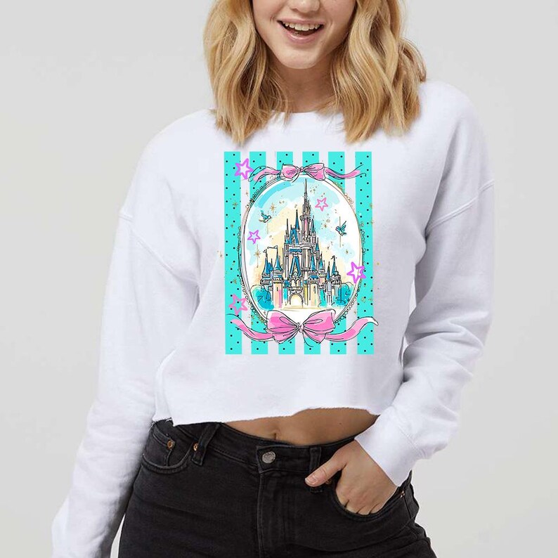 May include: White cropped sweatshirt featuring a colorful illustration of a castle within a teal and pink design. The design includes pink bows, stars, and a striped background. The sweatshirt is a casual, everyday wear item.