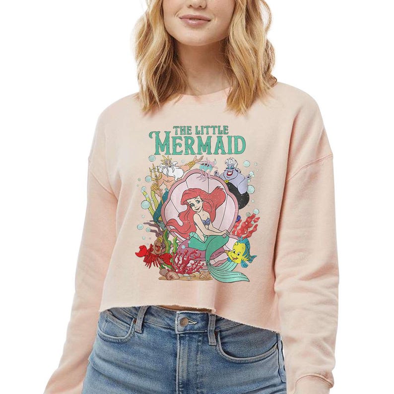 May include: A light peach-coloured cropped sweatshirt featuring a graphic print of The Little Mermaid characters including Ariel, King Triton, Ursula, Sebastian, and Flounder. The design includes the text THE LITTLE MERMAID in a teal font.