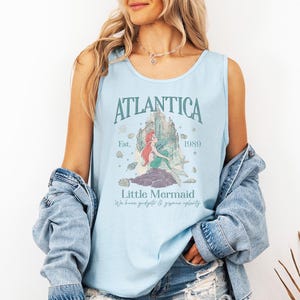 Op de afbeelding: Lichtblauwe tanktop met het woord "Atlantica" en een afbeelding van een zeemeermin. De tekst "Little Mermaid" en "We have gadgets & gizmos aplenty" staan ook op de top. De tanktop is lichtblauw.