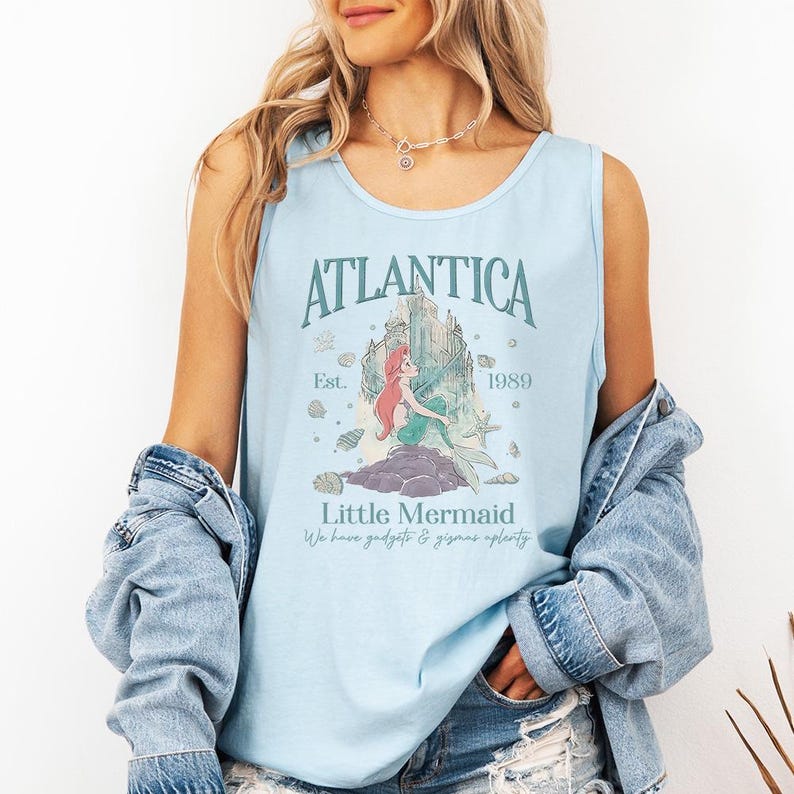Op de afbeelding: Lichtblauwe tanktop met het woord "Atlantica" en een afbeelding van een zeemeermin. De tekst "Little Mermaid" en "We have gadgets & gizmos aplenty" staan ook op de top. De tanktop is lichtblauw.