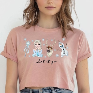 May include: A cropped, dusty rose t-shirt featuring a graphic of characters from the movie Frozen, including Elsa, Olaf, and Sven, with the phrase "Let it go" in a script font. The design includes snowflakes and a snowflake wand.